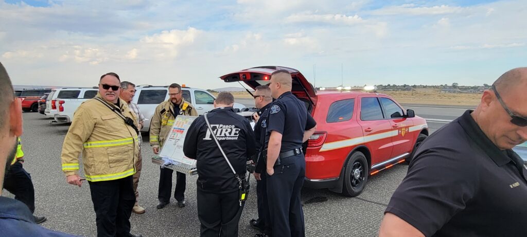 Emergency responders gather around a vehicle, discussing operations near an airport. A red fire department SUV is parked nearby.