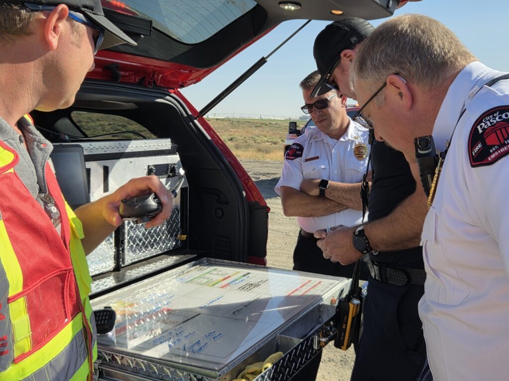 Emergency responders examine equipment in the back of a red vehicle, discussing plans and analyzing data in a desert setting.