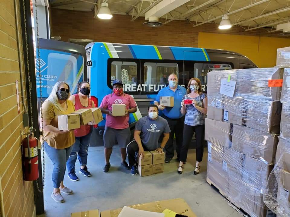 Volunteers at a community center hold boxes of supplies, standing near a van and stacked donations, showcasing their support for local youth programs.