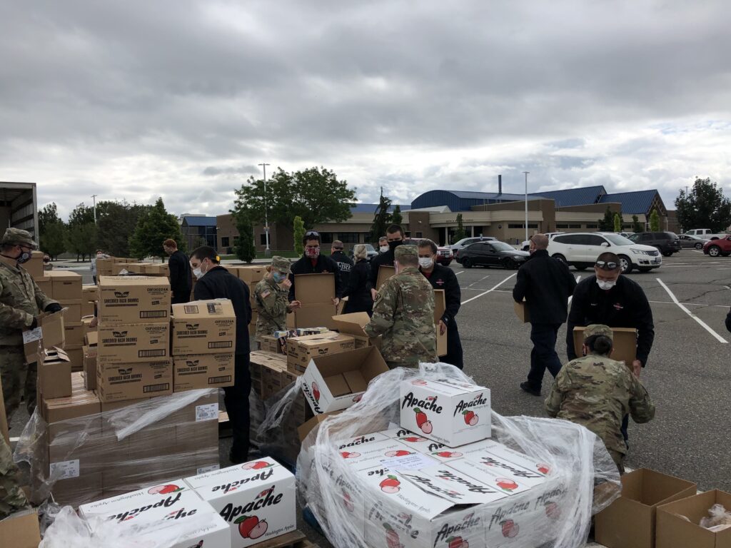Volunteers and military personnel distribute food boxes outdoors, with cardboard boxes stacked around them on a cloudy day.
